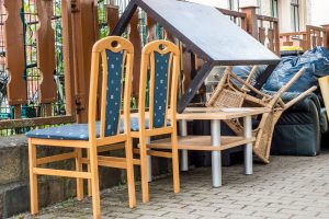 Zwei Holzstühle und verschiedene weggeworfene Haushaltsgegenstände, darunter ein kleiner Tisch und Weidenkörbe, wurden während einer Entrümpelung neben einem Metallzaun auf einem Gehweg aufgestapelt.